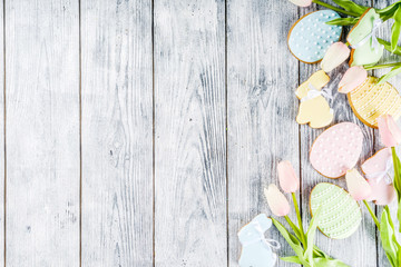 Easter greeting card background with pastel colored eggs and homemade cookies shaped in eggs and bunnies rabbits. With a basket, tulips, rustic wooden table, copy space top view banner