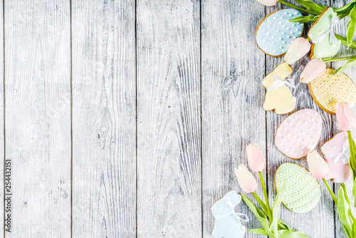 Easter greeting card background with pastel colored eggs and homemade cookies shaped in eggs and bunnies rabbits. With a basket, tulips, rustic wooden table, copy space top view banner