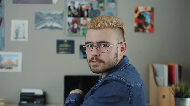Portrait of caucasian handsome man with glasses and stylish haircut yellow hair sitting at home office desk turn back and talking to camera. Speaks motivational speech.