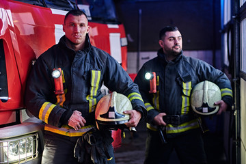 Two firemen wearing protective uniform standing next to a fire engine in a garage of a fire department.