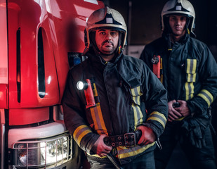 Two firemen wearing protective uniform standing next to a fire engine in a garage of a fire...