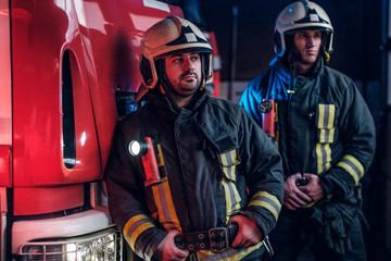 Two firemen in the fire station garage, standing next to a fire truck and looking sideways