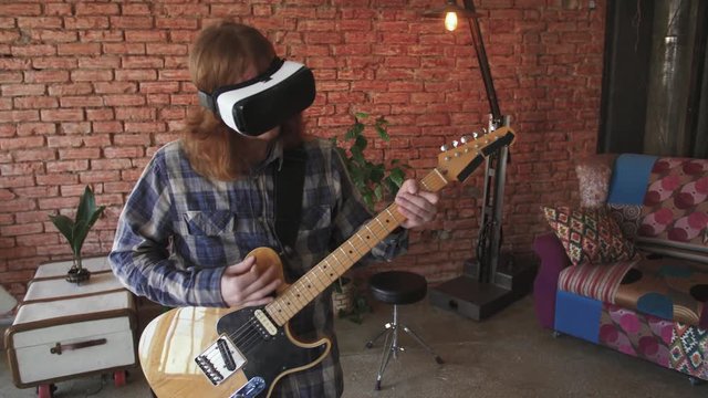 Portrait of young redhead man musician playing electric guitar in VR glasses at home, slow motion