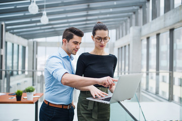 Two young businesspeople with laptop standing in office, talking.