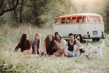 A group of young friends with a dog sitting on grass on a roadtrip through countryside.