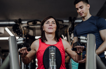 A female senior with a young trainer doing strength workout exercise in gym.