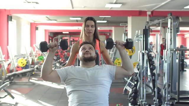A caucasian man with a beard performs an exercise in the gym bench dumbbells sitting with the help of a beautiful girl trainer