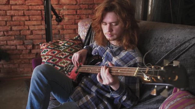 Portrait of young man musician with long red hair playing bass guitar at home, sitting on sofa