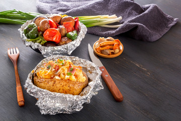 Baked potatoes with bacon, onions and baked vegetables in foil - tomatoes, eggplants, peppers on a gray wooden table.