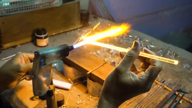 The man's hand is holding the glass stick to the fire to melt and mold, craft art from the blown glass.