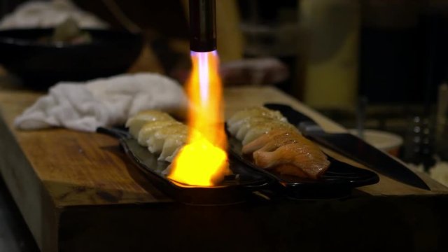 Asian cheff burns a slice of raw fish with blowtorch for make a sushi caramelize. Man cooking in a japanese restaurant. Searing and burning fresh sashimi on a kitchen background, Japan. -Dan