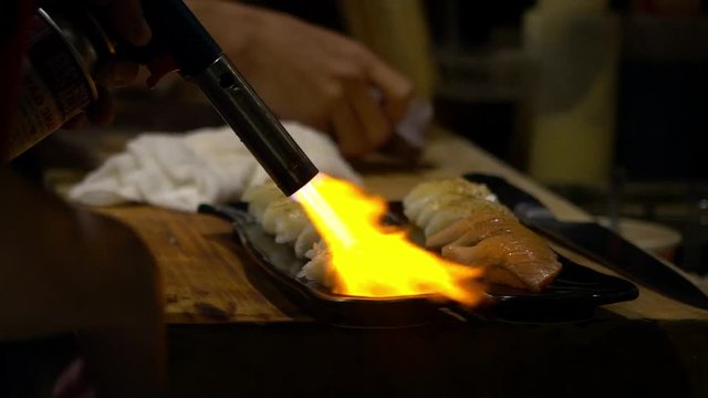 Asian cheff burns a slice of raw fish with blowtorch for make a sushi caramelize. Man cooking in a japanese restaurant. Searing and burning fresh sashimi on a kitchen background, Japan. -Dan