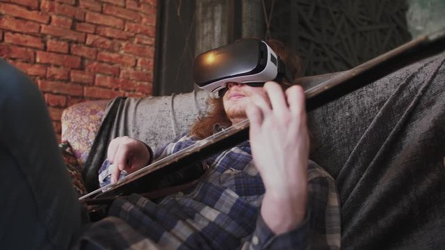 Young redhead man musician playing bass guitar in VR glasses at home, sitting on sofa, close up