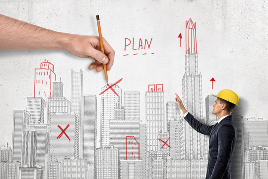 Side view of businessman in hard hat beside wall with city plan on it and big male's hand on other side crossing out some landmarks with pencil.