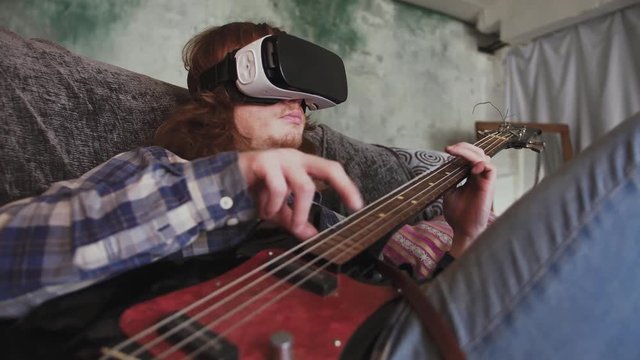 Young redhead man musician playing bass guitar in VR glasses at home, sitting on sofa
