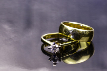 Gold antique jewelry on the silver metal surface with reflections.Simple marriage ring put on the golden engagement ring with diamond gem. On the mirror surface with the reflections