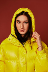 Portrait of brunette young woman wearing oversized yellow down jacket with hood on. Studio close up shot.