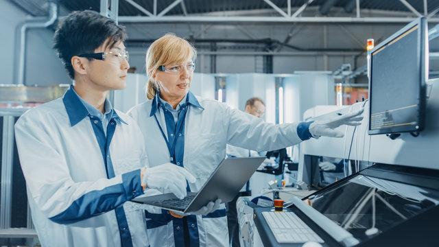 On High Tech Factory Worker Load Reels Into Pick And Place Machine, Engineers Use Computer For Programming Machinery For Printed Circuit Board Assembly Line. Production Of PCB With SMT Machinery.