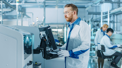 Obraz premium On High-Tech Factory Male Engineer Uses Computer for Programing Pick and Place Electronic Machinery for Printed Circuit Board Surface Mount Assembly Line. Production of PCB with SMT Machinery.