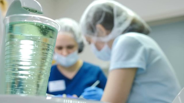 Shot of plastic glass with water on first plan and blurred dentist and assistant making dental procedure on background. Closeup shot of dentist hands and young woman with cofferdam in mouth. Doctor