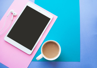 Flat lay of white tablet, earphones and a cup of coffee on blue, purple and pink background with...
