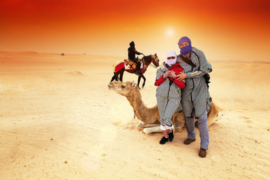 Tourists in the Sahara desert near to camel.In the background is rider on the horse, unrecognizable person.Sunset light, lens flare