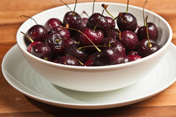 Top, front view , close distance of a white ceramic bowl of ripe, local, raw cherries with stems, rinsed on  white ceramic saucer, on a wood tabl