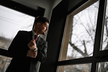 A handsome man in a black suit and a red tie posing at the big window. Business portrait of a man.