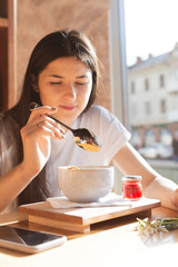 beautiful brunette girl wearing a white shirt sitting at breakfast table in bright Pastries near a large window