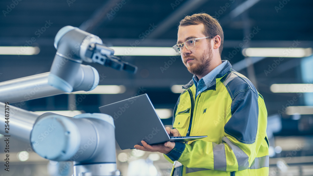Poster At the Factory: Automation Engineer Uses Laptop for Programming Robotic Arm. New Era in Automatic Manufacturing Industry.