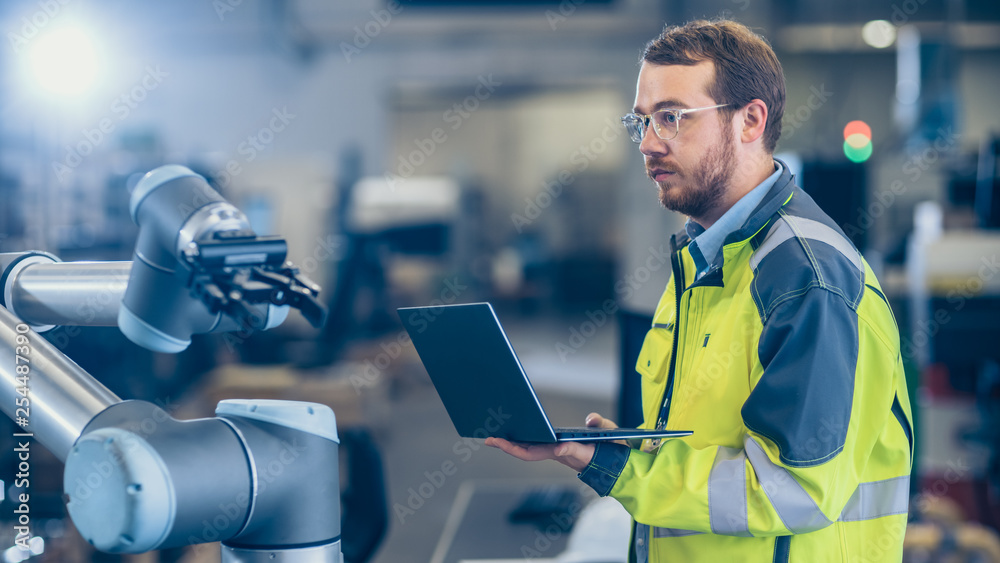 Fototapete At the Factory: Automation Engineer Uses Laptop for Programming Robotic Arm. New Era in Automatic Manufacturing Industry.