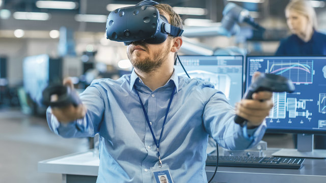 Portrait Shot of the Industrial Engineer Wearing Virtual Reality Headset and Using Controllers, ready to Work. In the Background Manufacturing Plant and Monitors. - Powered by Adobe