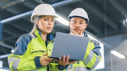 Low Angle Shot of Male and Female Industrial Engineers Work on a Manufacturing Plant, They Discuss Project while Using Laptop.In the Background Industrial Manufacturing Plant.