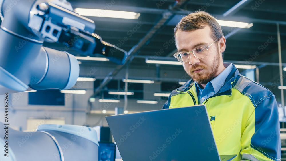 Poster At the Factory: Automation Engineer Uses Laptop for Programming Robotic Arm. New Era in Automatic Manufacturing Industry.