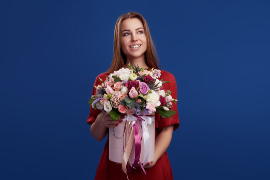 Wow effect. Wonderful flowers on Womens Day. Tender smiling brunet woman poses with bouquet of colorful flowers in hat box, looks happily, isolated over blue background wall. Space for text - Powered by Adobe