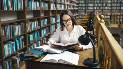 Cheerful dark haired student smiling while sitting at desktop with many studying books for exam preparation