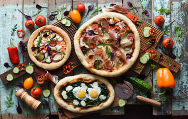 Homemade pizza party. Flatlay of puffy rustic pizzas with pancetta, aubergines, tomatoes, spinach, eggs, bell peppers and arugula served with raw vegetables and herbs on shabby blue background