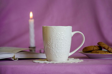 Still life of porcelain cup of tea with cookies, opened book and  burning candle. Romantic interior in lilac white colors. Close up, selective focus.