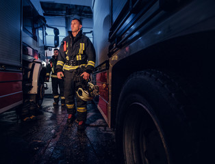 Obraz na płótnie Canvas Full-length portrait of two brave firemen in protective uniform walking between two fire engines in the garage of the fire station