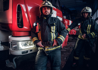 Two firemen wearing protective uniform standing next to a fire engine in a garage of a fire department.