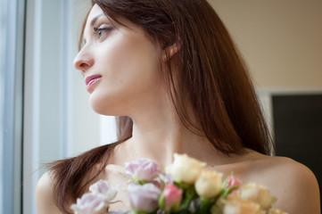 Naklejka premium Young woman with bouquet of the flowers looking through the window with tenderness early in the morning at home