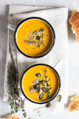 Two portions of fresh homemade pumpkin creamy soup served with liver and seeds in a white enamel mugs on a light grey concrete background. View from above, flat lay.