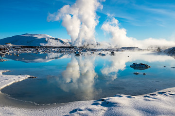 Steam from a geothermal plant set against a bright blue sky and reflected in a lake beside the Blue...