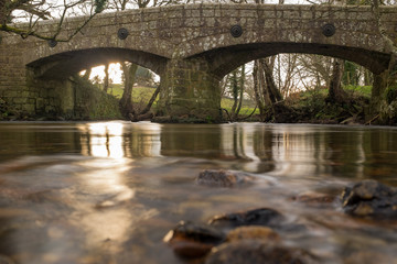 Fototapeta premium The River Teign glides under an Ancient stone bridge just outside Chagford in Devon, England, out of focus rocks in the foreground pointing towards the bridge