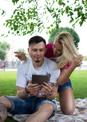 Cheerful couple using digital tablet in the park sitting on a green lawn. Sexy girl and boyfriend watch interesting information on the Internet. Conceptual photo