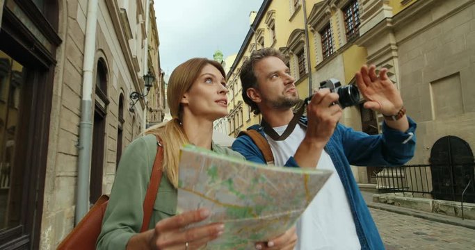 Caucasian handsome man with camera and pretty blonde woman with map walking the street together, taking photos and talking.