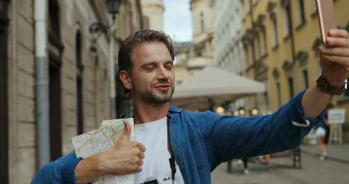 Close up of the good looking young man tourist on the city street taking selfie photo with a map on the smartphone camera and giving his thumb up.