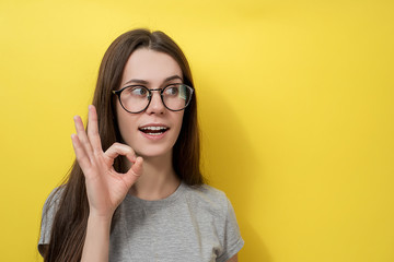 Optimistic girl in glasses shows excellent gesture, with amazed look, dressed in gray T-shirt, isolated over yellow background. Female demonstrates okay sign