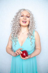 blonde curly girl in blue dress holding tasty big red apple and smile on white studio solid background