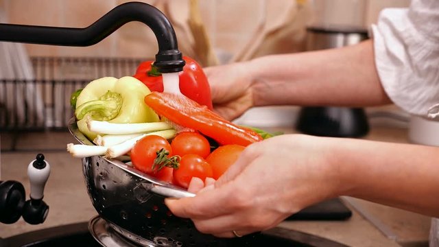 Woman hands washing lots of vegetables at the kitchen sink - holding a strainer under the tap water flowing, slow motion, static camera